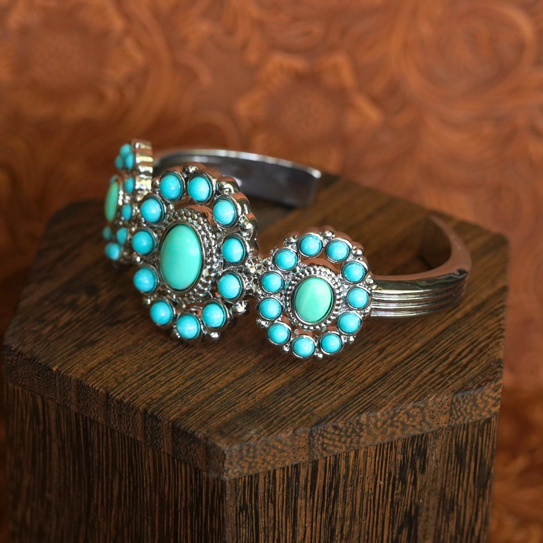 The Nellie Cuff, a silver Western bracelet featuring three circular turquoise stone clusters, is displayed on a textured wooden block against a brown patterned background.