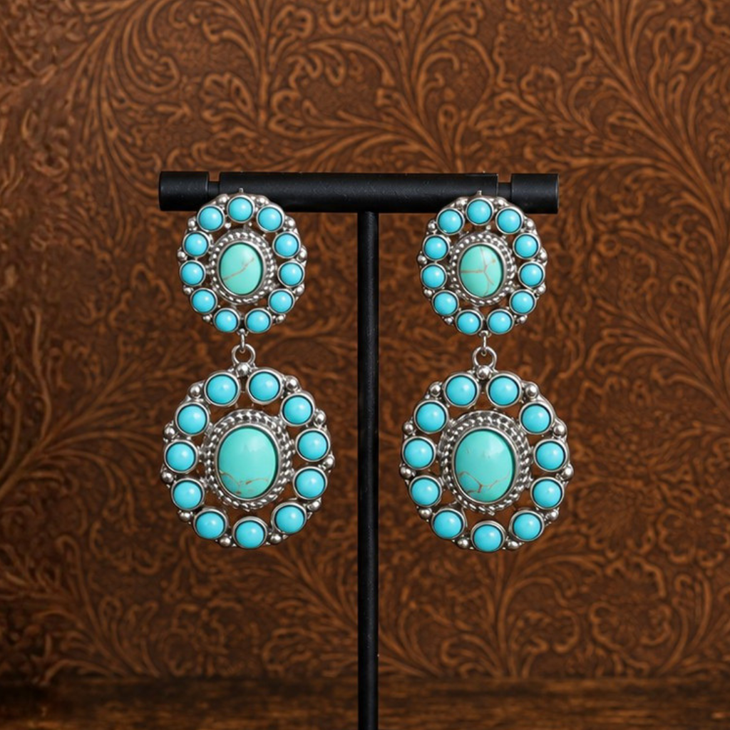 The Nellie Drop Earrings feature large, circular turquoise stones set in silver and are displayed on a stand against a brown, ornate background—perfect statement earrings for adding a bold, Western-inspired touch to any look.