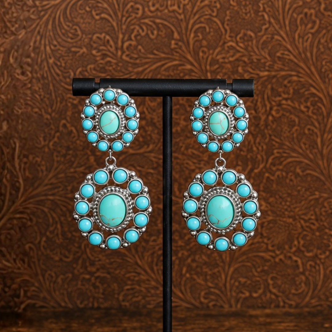 The Nellie Drop Earrings feature large, circular turquoise stones set in silver and are displayed on a stand against a brown, ornate background—perfect statement earrings for adding a bold, Western-inspired touch to any look.