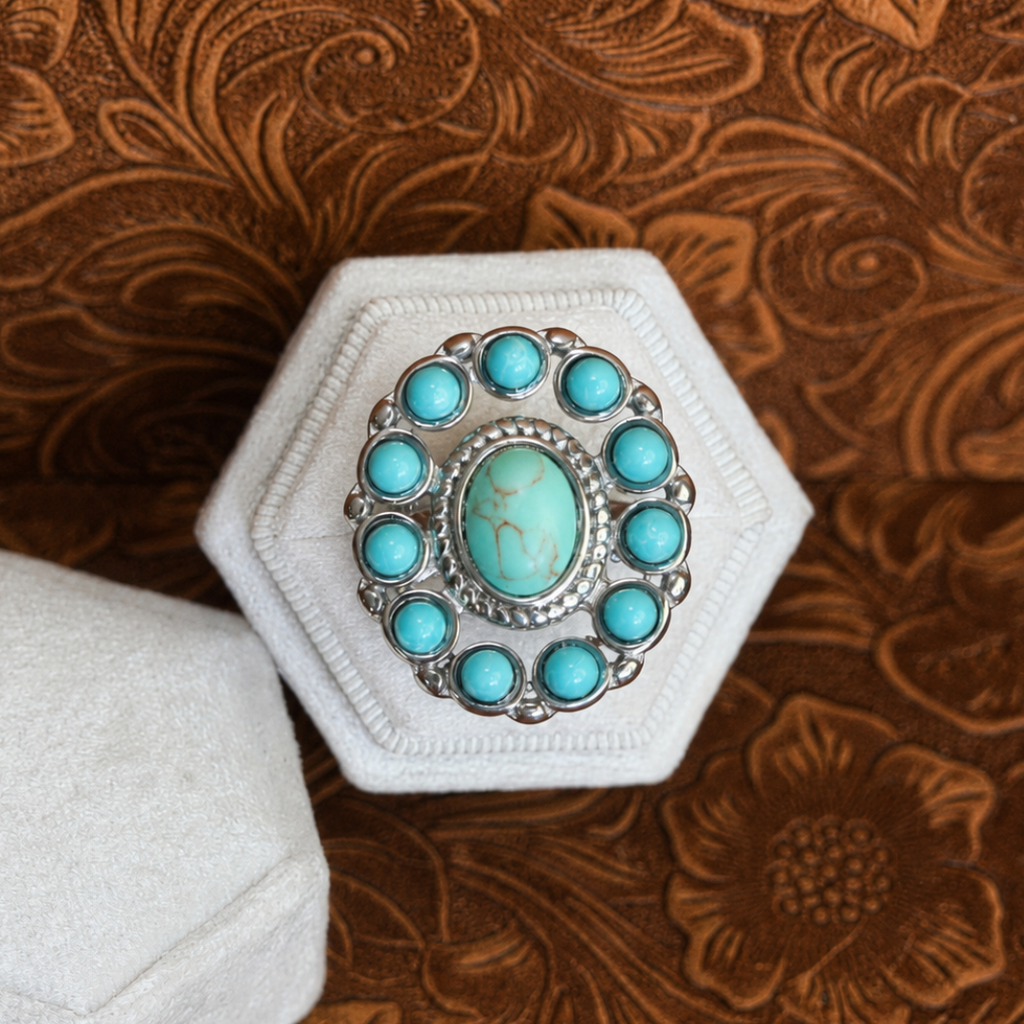 The Nellie Ring, a striking turquoise piece showcasing a large oval turquoise stone at its center surrounded by smaller round turquoise stones, rests in a white hexagonal jewelry box atop brown floral fabric—a stunning example of American West jewelry.