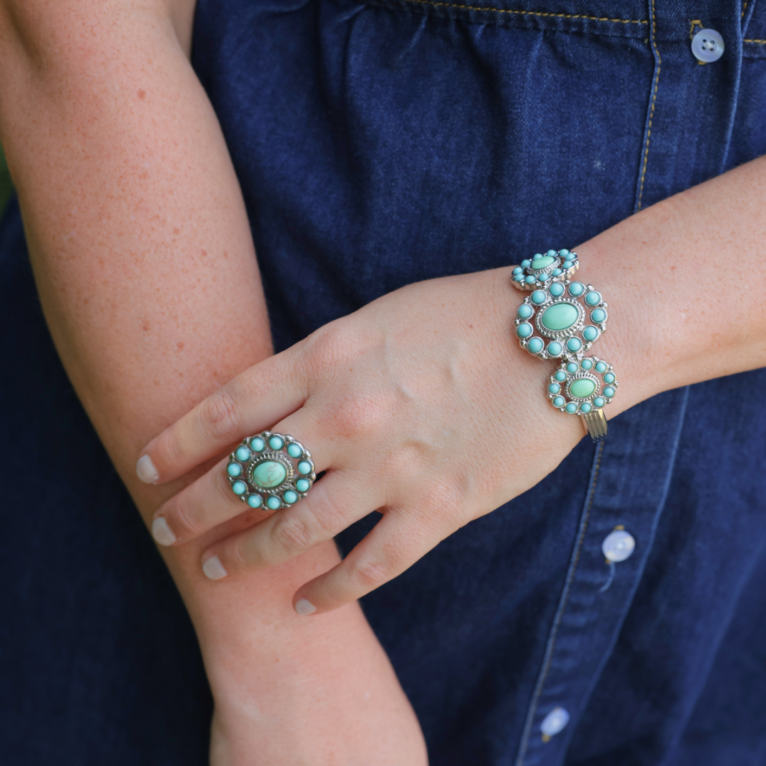 Wearing a blue denim dress, a person showcases American West jewelry style with a turquoise stone bracelet and The Nellie Ring—a bold Western statement piece—on their left hand.