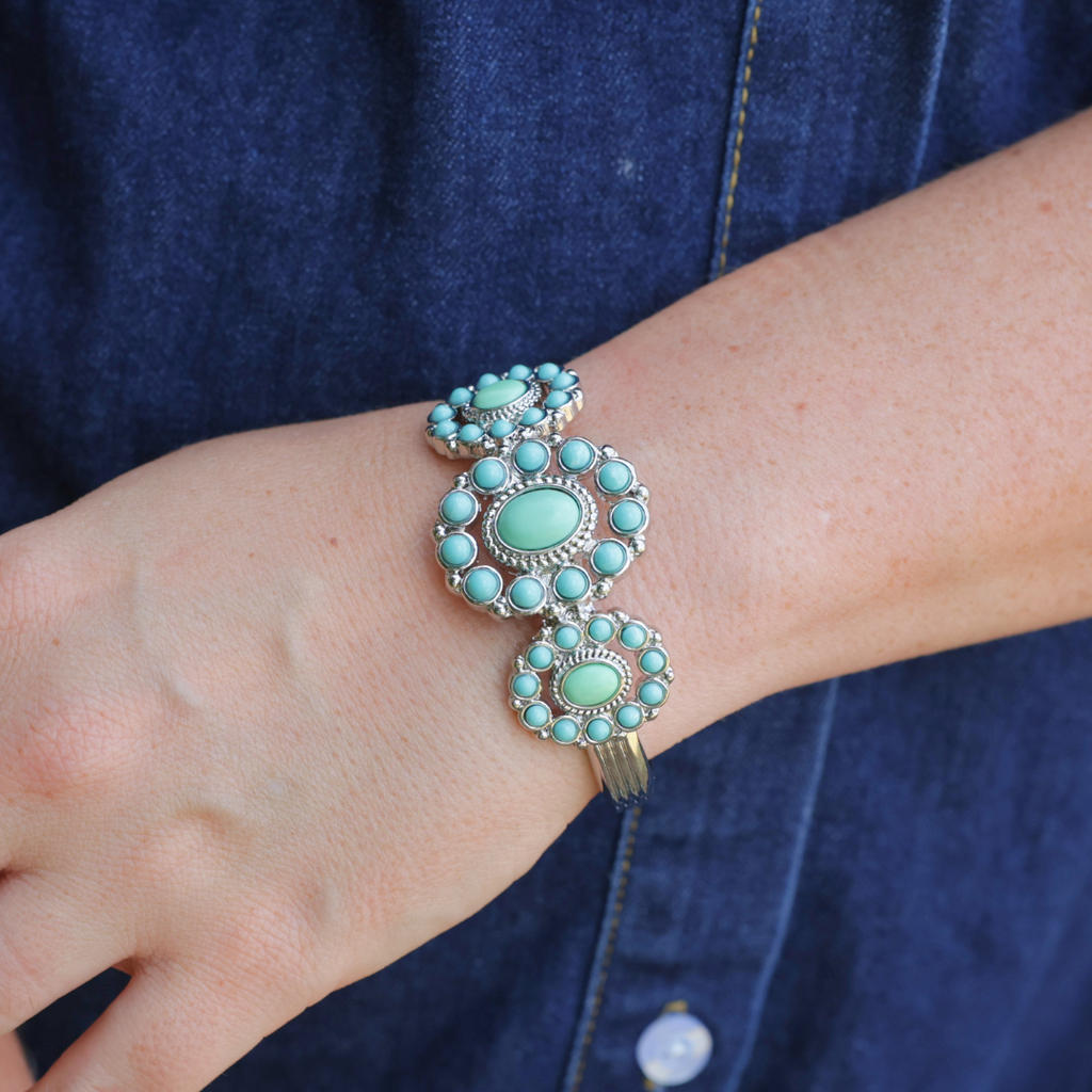 A hand wearing The Nellie Cuff—an elegant turquoise bracelet featuring three large floral clusters each encircled by small turquoise stones—rests against blue denim fabric, showcasing a heritage jewelry piece that brings classic Western charm to any outfit.
