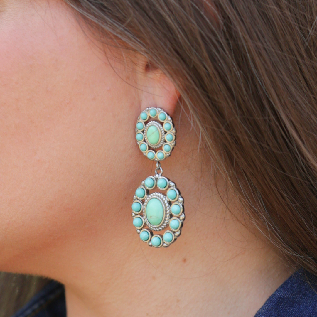 A close-up of a person with long brown hair wearing The Nellie Drop Earrings—bold, silver statement earrings featuring oval-shaped turquoise stones. Dressed in a blue top, the wearer highlights the striking beauty of these Western-inspired earrings.