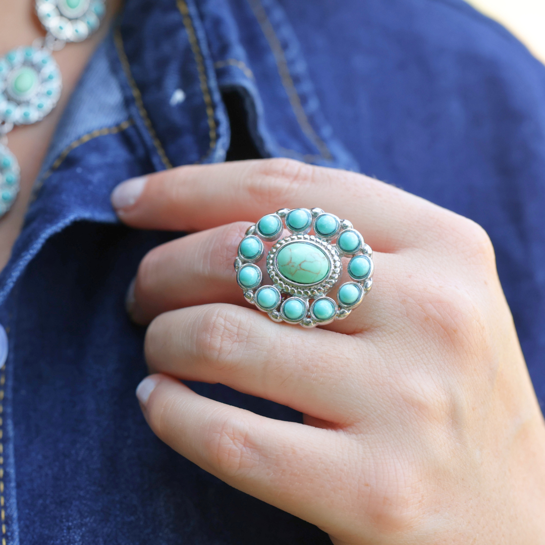 A hand adorned with The Nellie Ring—a large turquoise piece in classic American West jewelry style—rests against a blue denim shirt, complemented by a matching turquoise necklace.