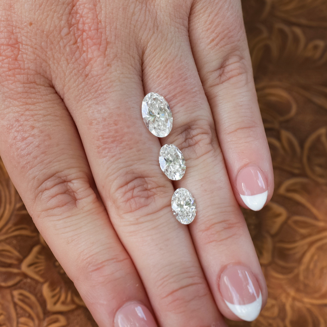 A hand with a French manicure displays three 1.5 ct. Oval Cut Lab Grown Diamonds, each IGI Certified with VS1 clarity, arranged in a row on top of the middle finger against a brown, patterned background.