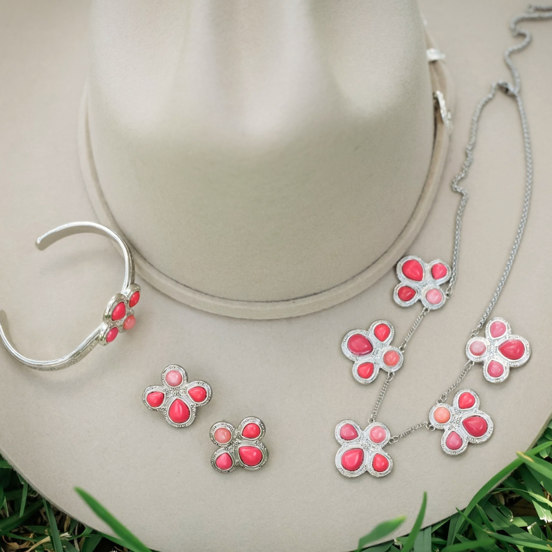 The Prairie Rose silver breast cancer awareness jewelry set, featuring flower-shaped pendants with pink stone inlays, including a cuff bracelet, earrings, and a Western style necklace resting on a beige felt hat.