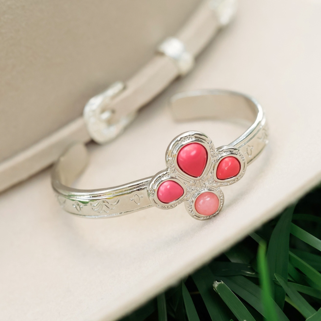 The Prairie Rose Cuff, a silver bracelet featuring a flower-shaped setting with four pink stones, rests on a beige hat as a symbol of Breast Cancer Awareness.