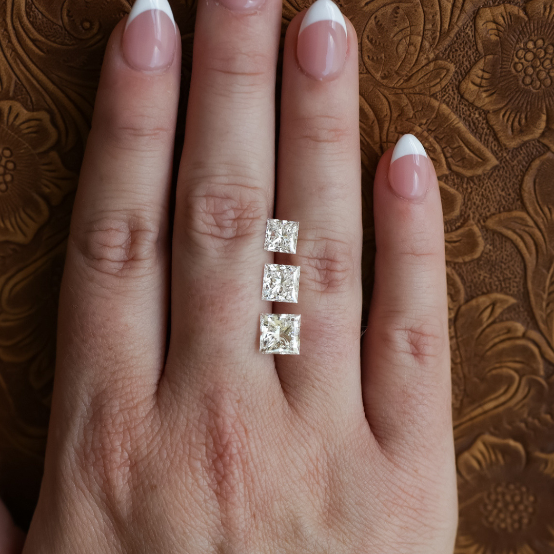 A hand with French-manicured nails rests on a brown, embossed surface. Three square-cut diamonds, including a stunning 1.5 ct. Princess Cut Lab Grown Diamond, are placed vertically on the middle finger.