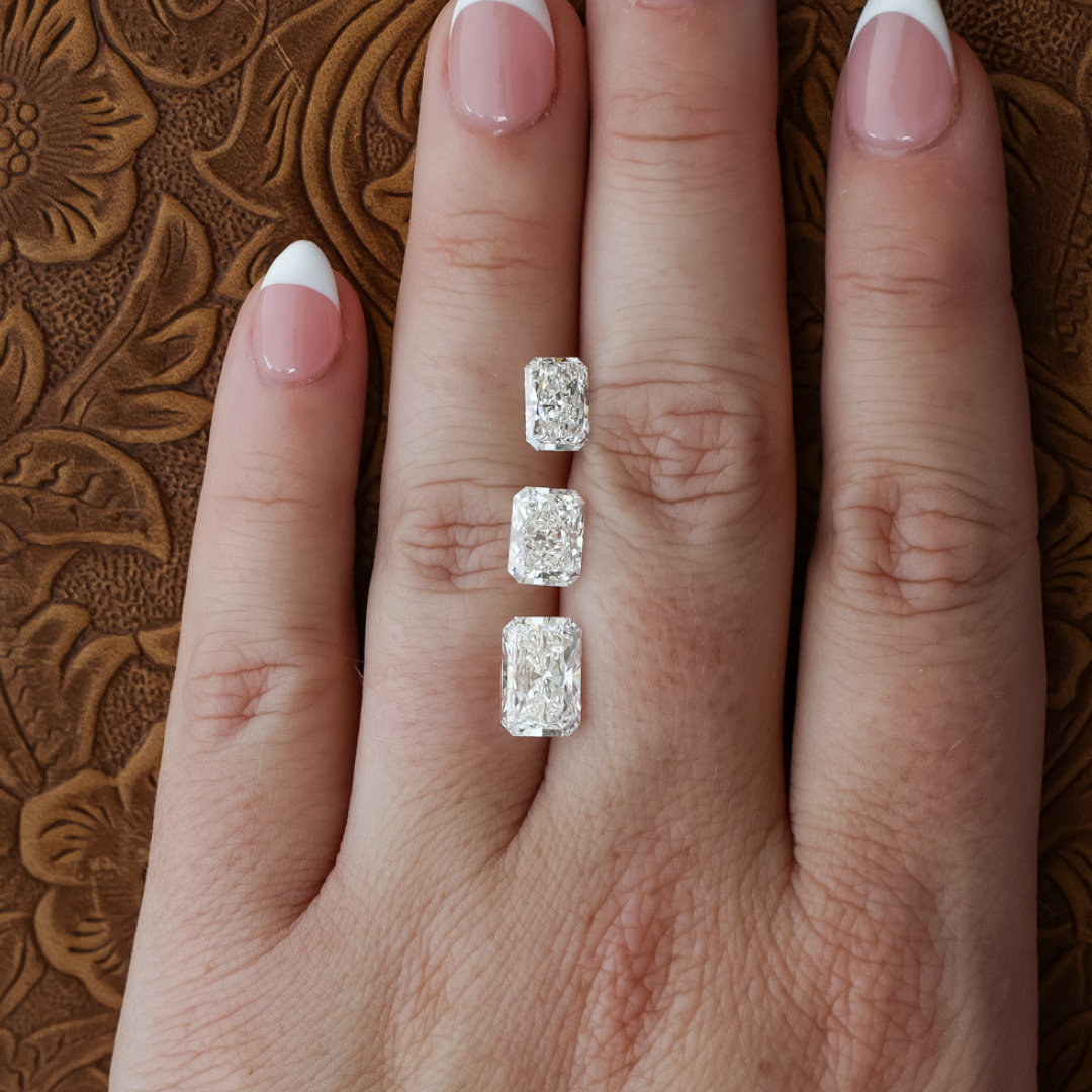 A hand with light pink and white French manicured nails showcases three rectangular Radiant Cut Lab Grown Diamond rings, featuring a stunning 1.5 ct. Radiant Cut Lab Grown Diamond on the middle finger, against a brown embossed background.