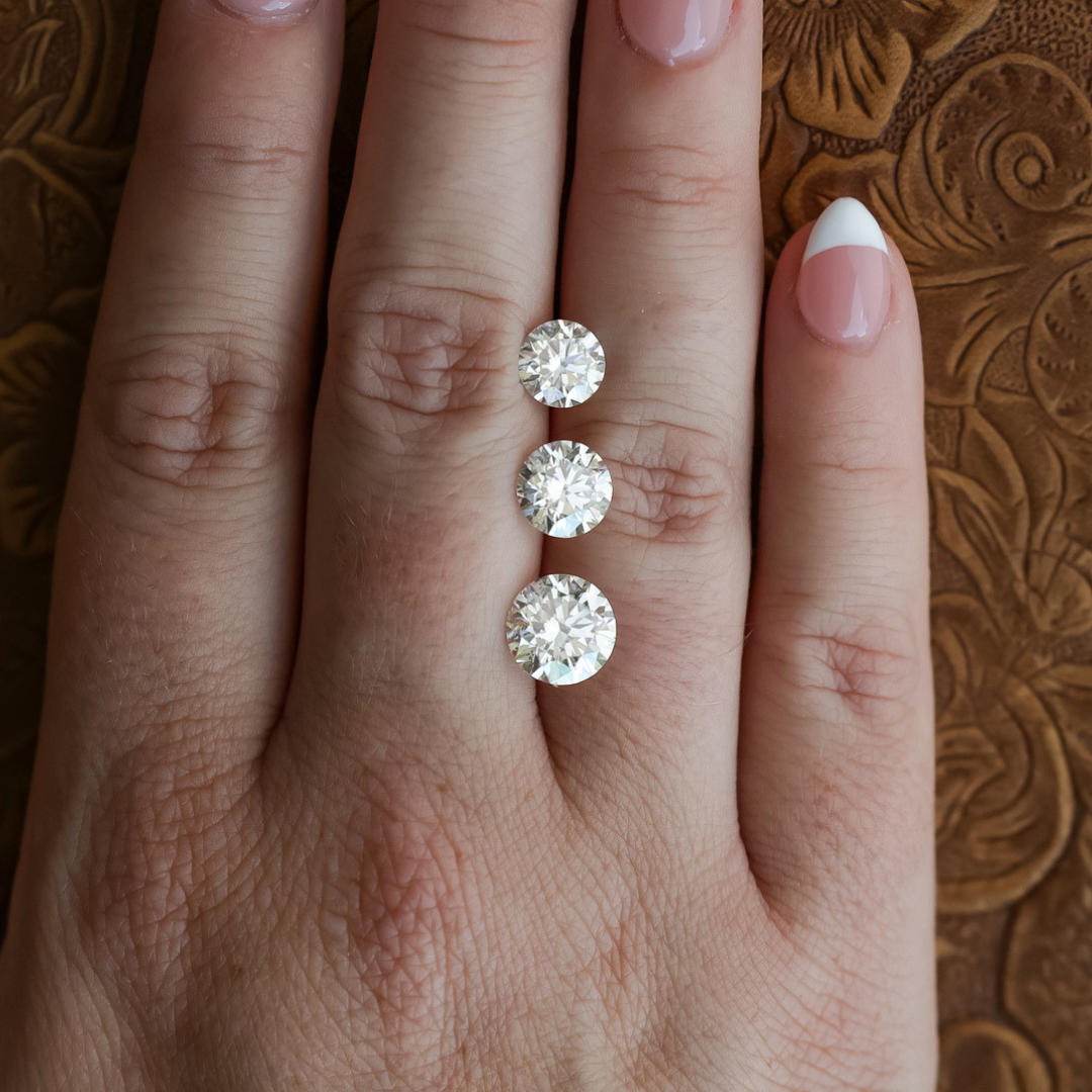 A hand with a French manicure rests on a textured brown surface, displaying three round diamonds of varying sizes—one of which is the 1.5 ct. Round Cut Lab Grown Diamond—placed along the middle finger.