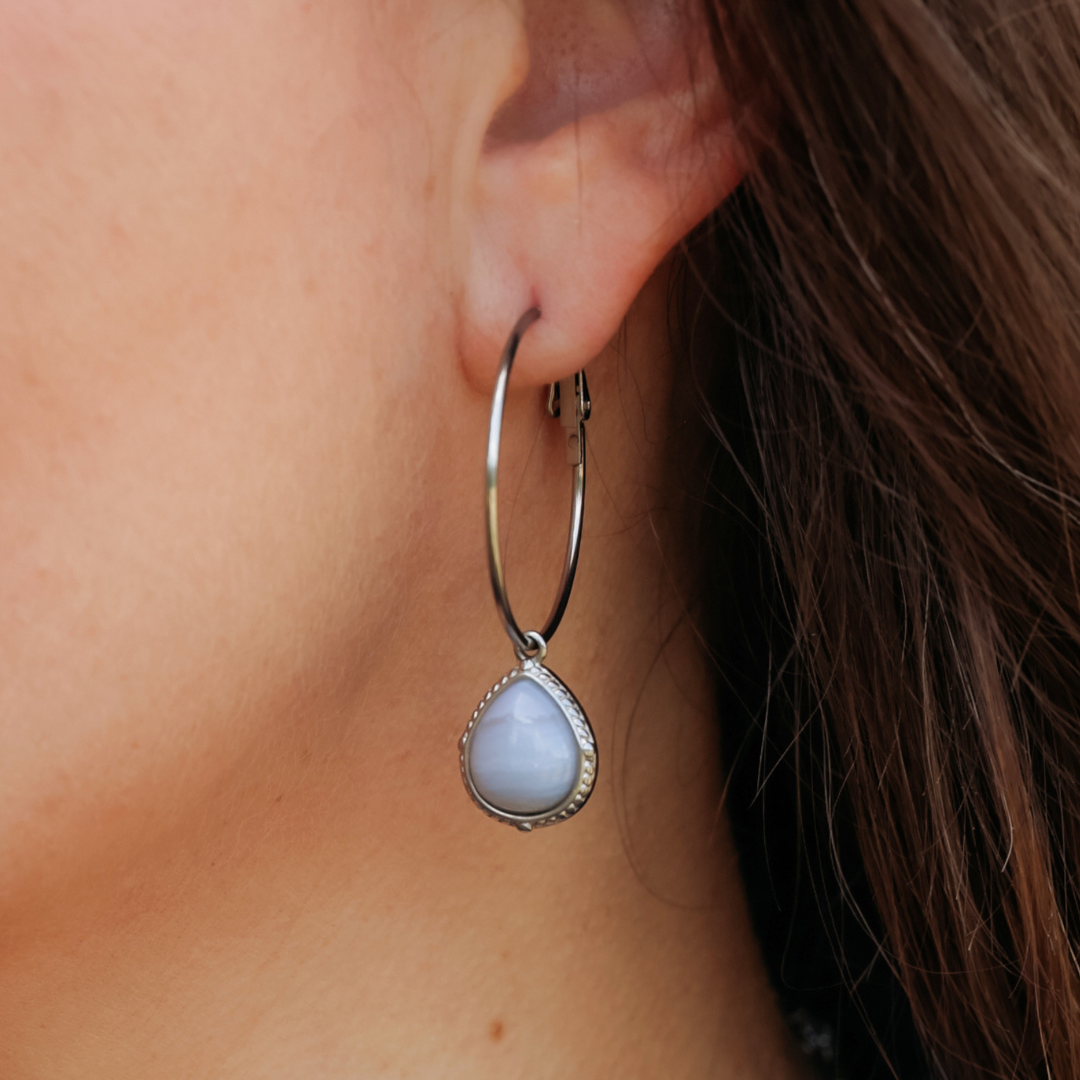 A close-up of a person’s ear wearing The Shay Hoops: handmade silver earrings featuring large western hoops adorned with a teardrop-shaped blue gemstone pendant.