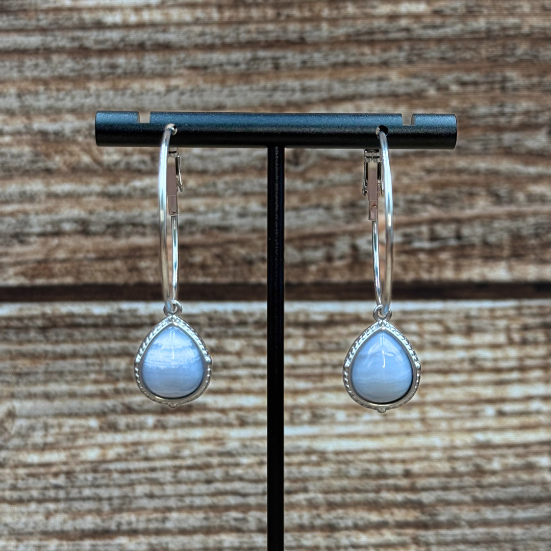 Introducing The Shay Hoops: Handcrafted earrings with silver hoops and light blue, teardrop-shaped gemstones suspended in the center. These striking turquoise hoop earrings are elegantly displayed on a stand against a wooden background.