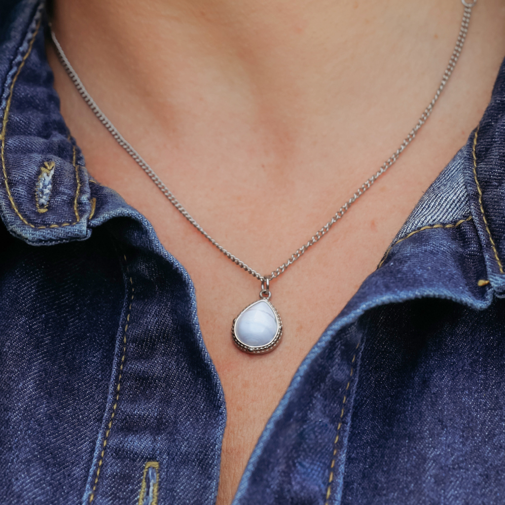 Close-up of a person wearing The Shay, a silver necklace featuring a teardrop-shaped blue lace agate pendant, stylishly paired with a blue denim shirt.