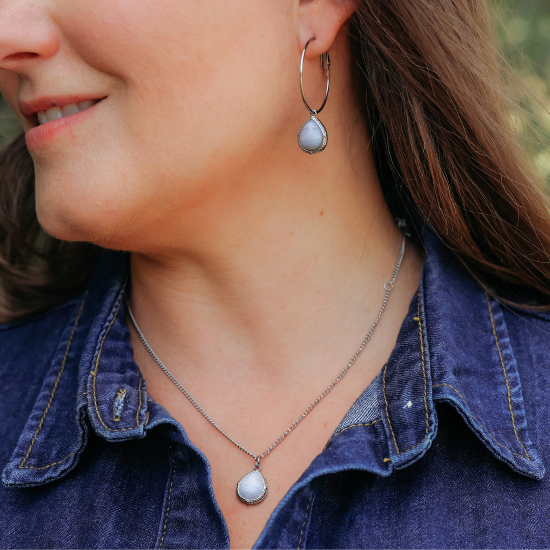 A woman wearing a denim shirt, The Shay Hoops—handcrafted silver earrings with teardrop-shaped pendants—and a matching silver necklace with a similar teardrop pendant. She is smiling slightly and facing to the left.