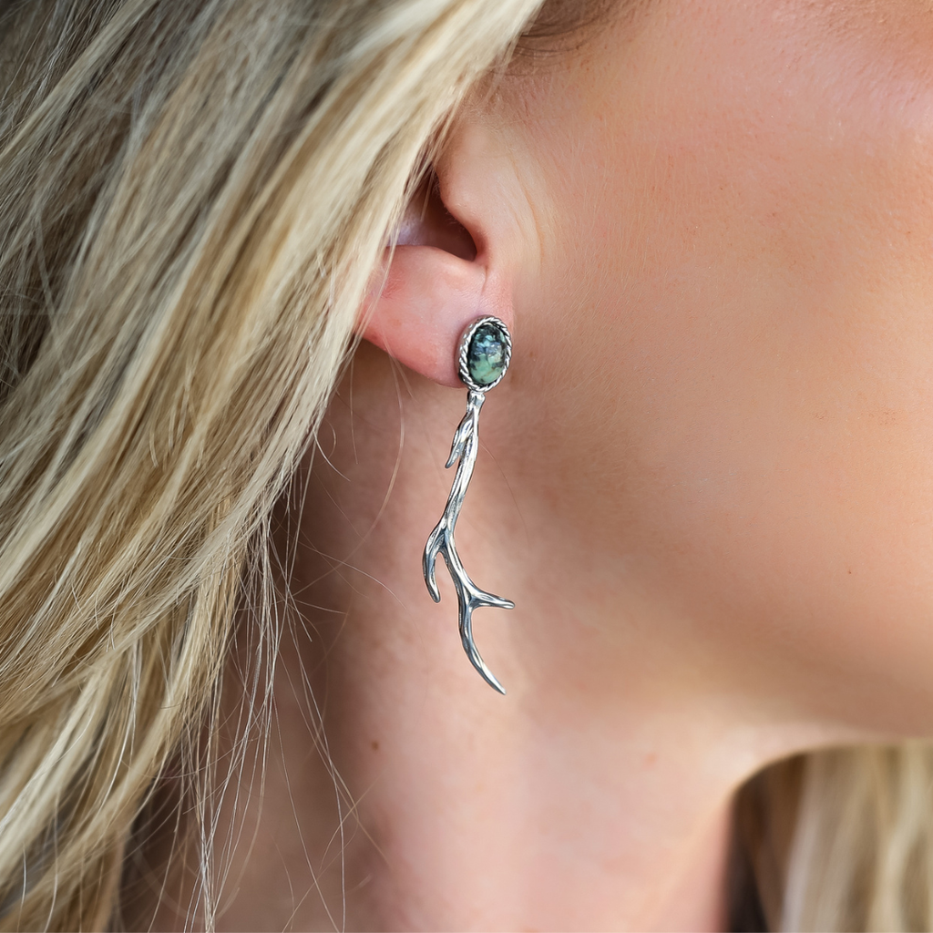 A close-up of a person wearing the Shed and Stone Drop Earrings—a silver, branch-shaped earring featuring a green gemstone at the top—set against their blonde hair and fair complexion. This handcrafted piece highlights the unique artistry of our Shed and Stone Drop Earrings collection, drawing inspiration from Western design elements.