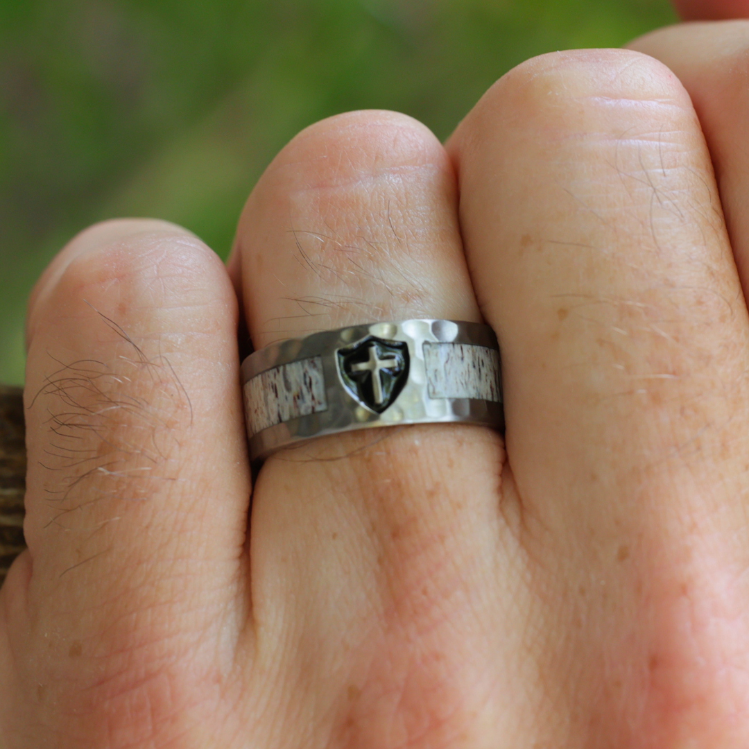 A close-up of a hand wearing The Shield of Faith, a silver ring engraved with a cross and shield design reminiscent of a Christian wedding band. The background is a soft, blurred green, likely grass or foliage.