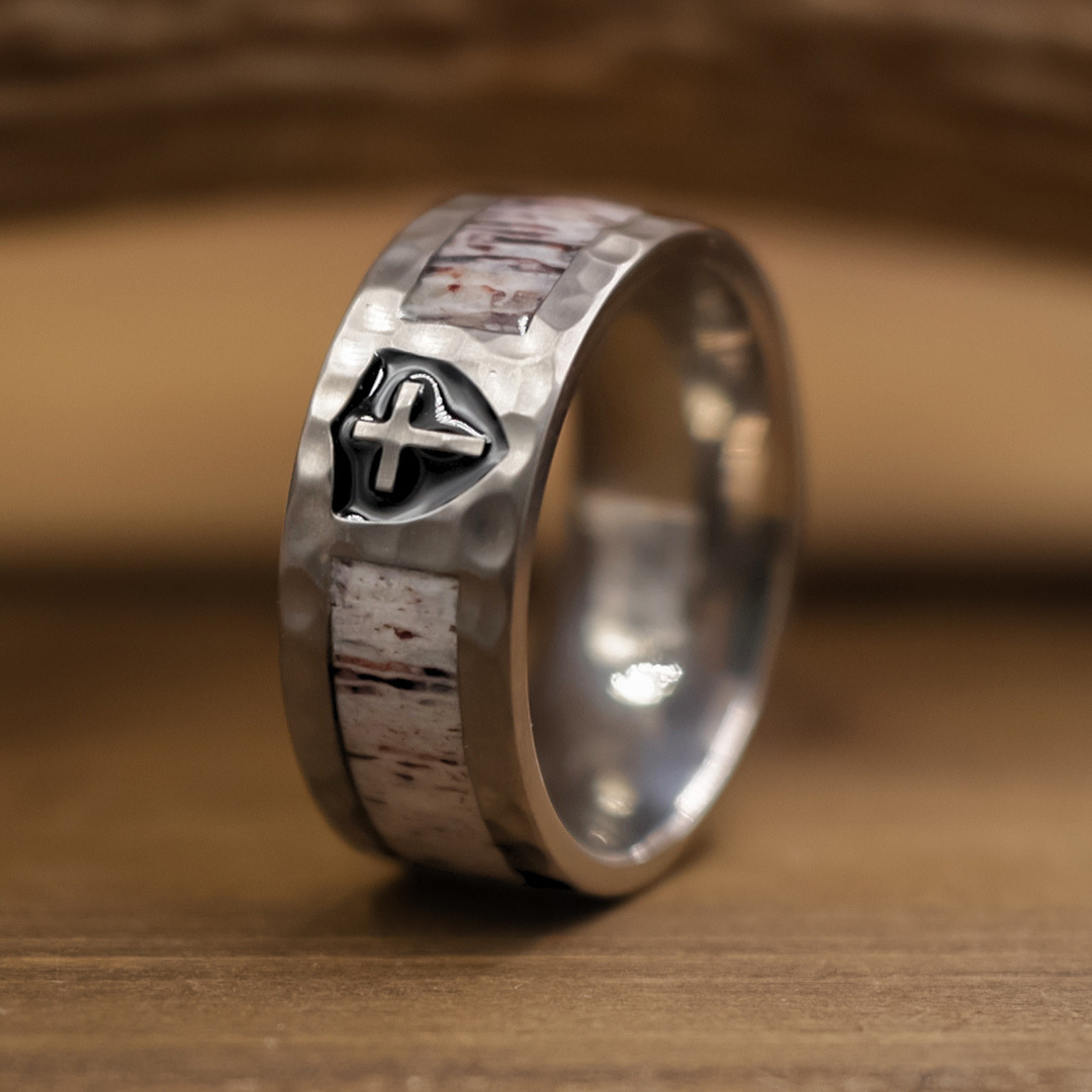 The Shield of Faith ring, featuring a hammered silver texture, white inlay, and a small cross emblem, is displayed upright on a wooden surface—an elegant Christian wedding band that beautifully highlights its distinctive hammered finish.