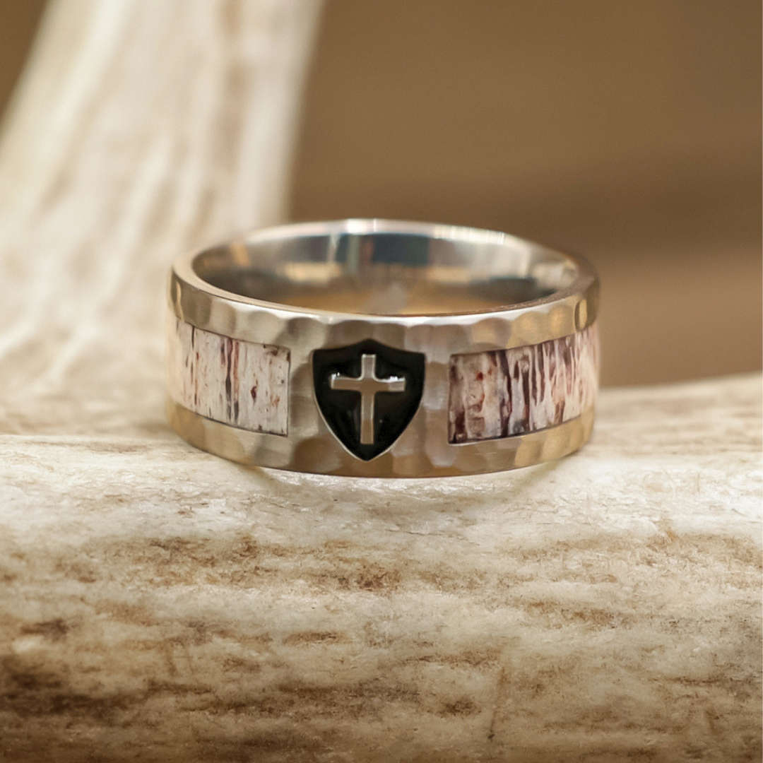 The Shield of Faith, a silver Christian wedding band adorned with a black shield and cross emblem, showcases rustic wood-like inlays and a subtle hammered finish as it rests on a textured, light-colored antler.