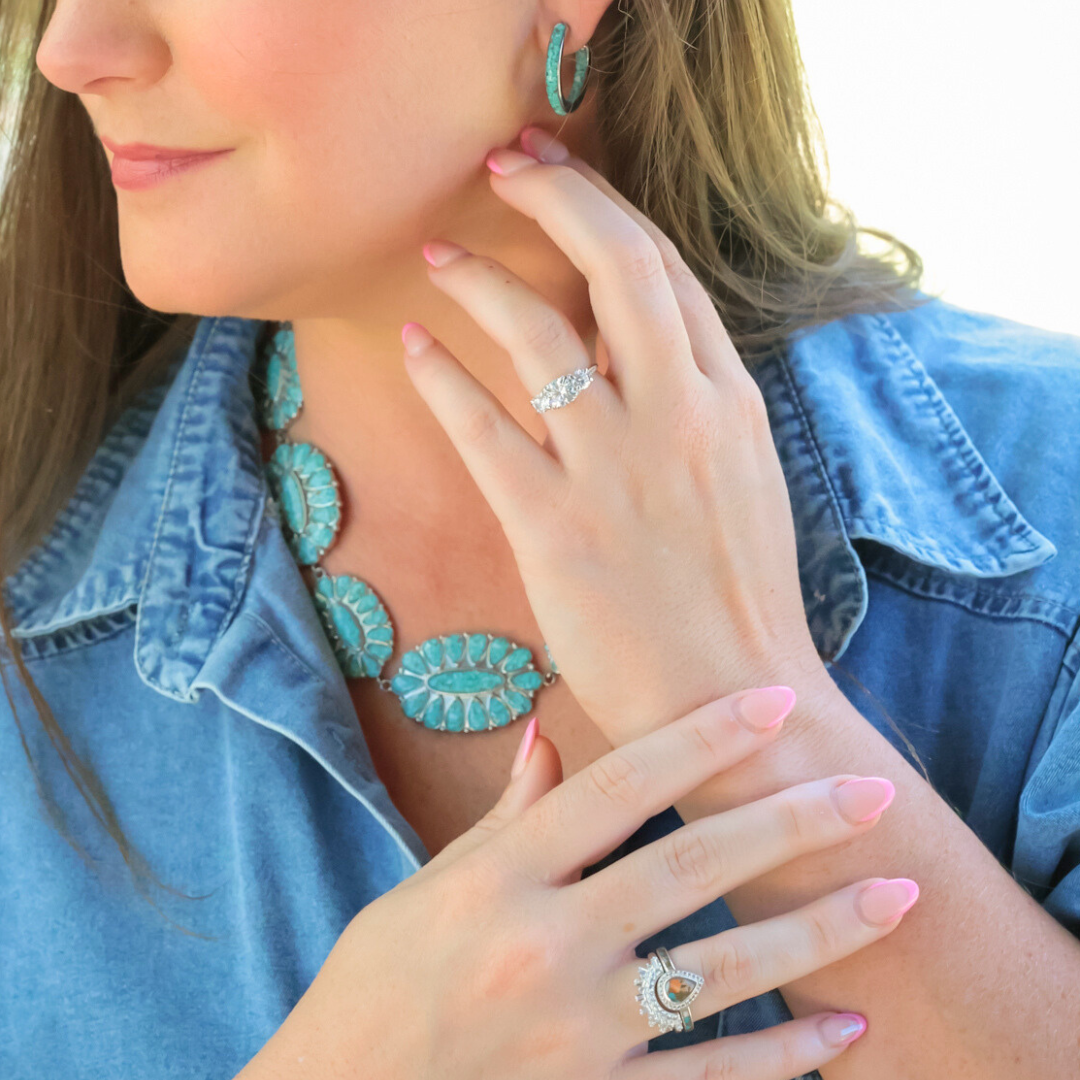 A woman with light pink nails wears turquoise jewelry, a blue denim shirt, and two rings, one with a large stone and another with a teardrop-shaped stone, touching her face and neck.