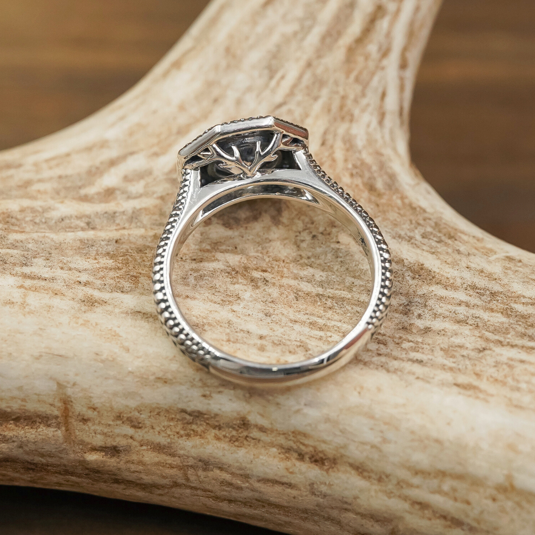 A silver engagement ring with intricate detailing is displayed upright on a textured antler, with a wooden background visible.