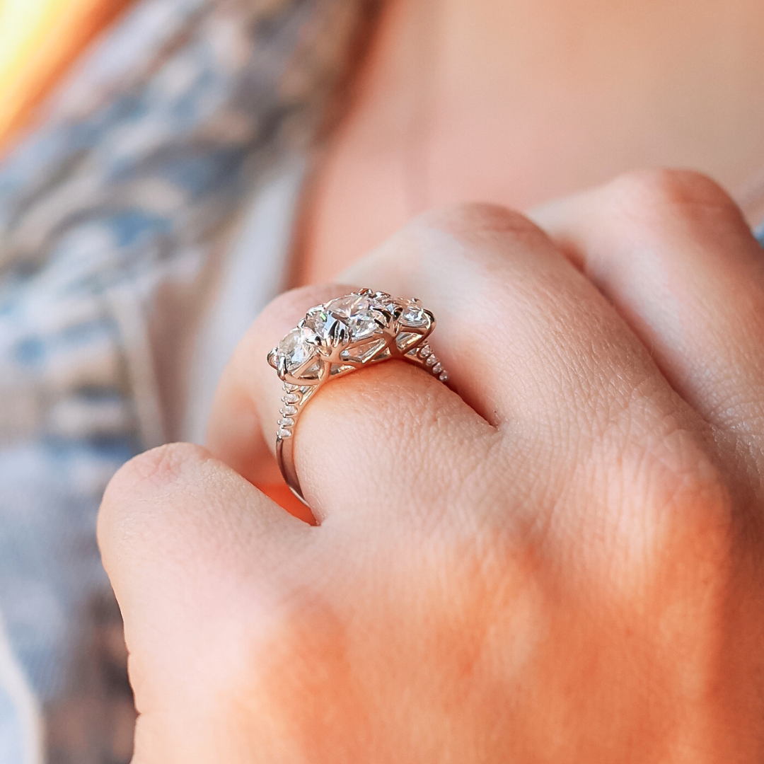 A close-up of a hand wearing a silver ring with three clear gemstones, with a blurred, casual patterned shirt in the background.