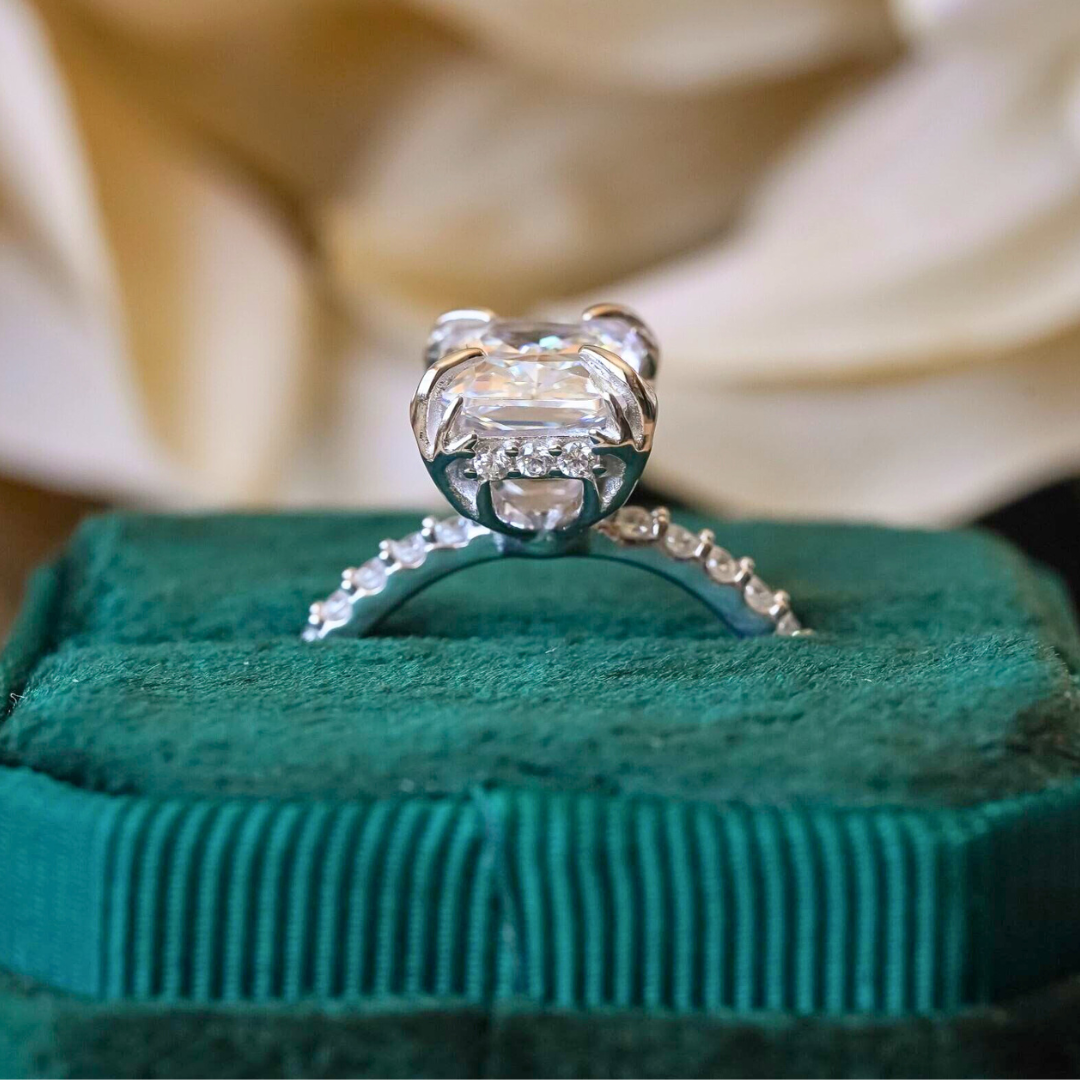 A diamond engagement ring with a decorative setting is displayed on a green velvet box, with a blurred white flower in the background.