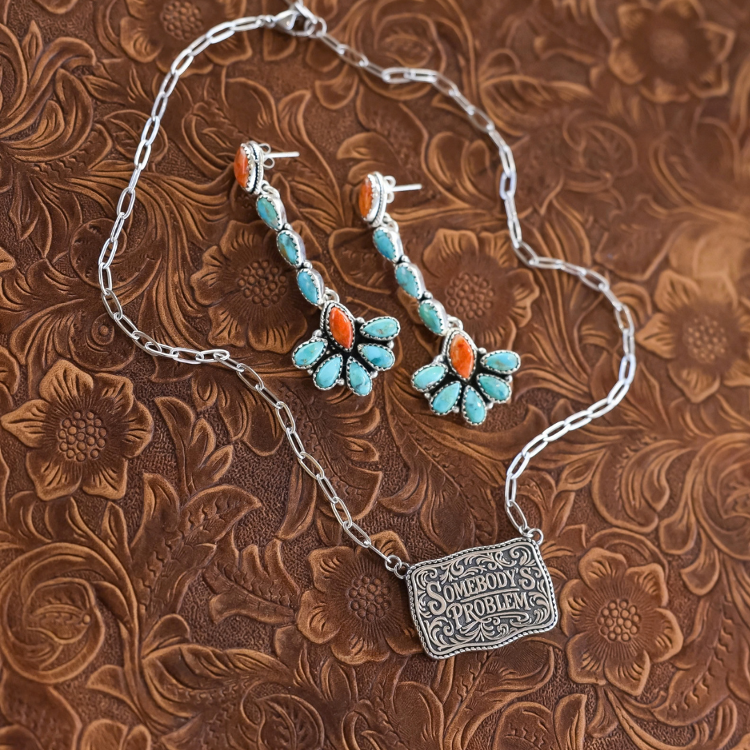 The Somebody’s Problem silver Western necklace features a rectangular pendant embossed with the words Somebodys Problem and matching drop earrings with turquoise and coral-colored stones.