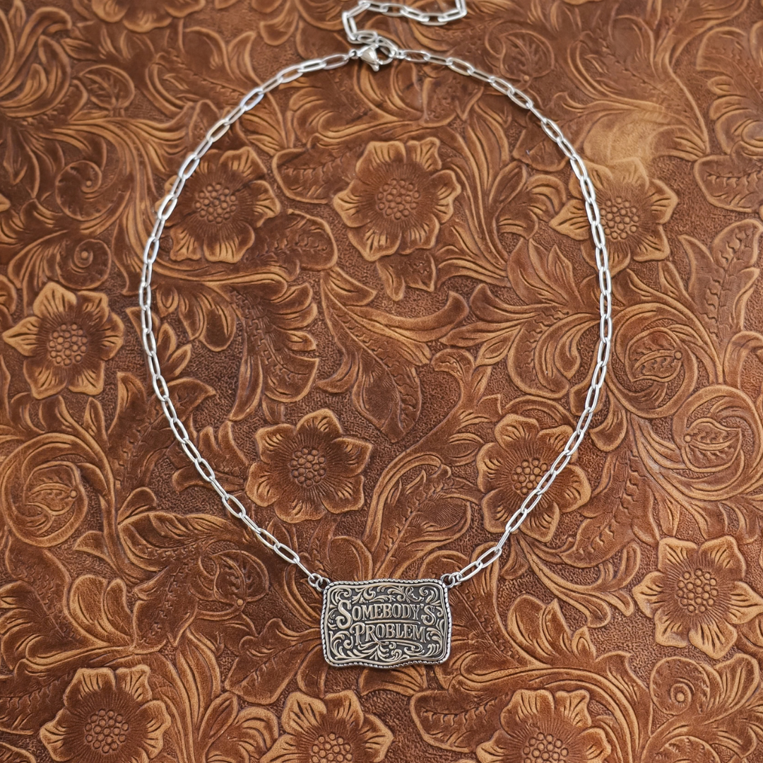 The Somebody’s Problem is a silver statement necklace featuring a rectangular pendant engraved with the words Somebodys Problem in a decorative Western font.