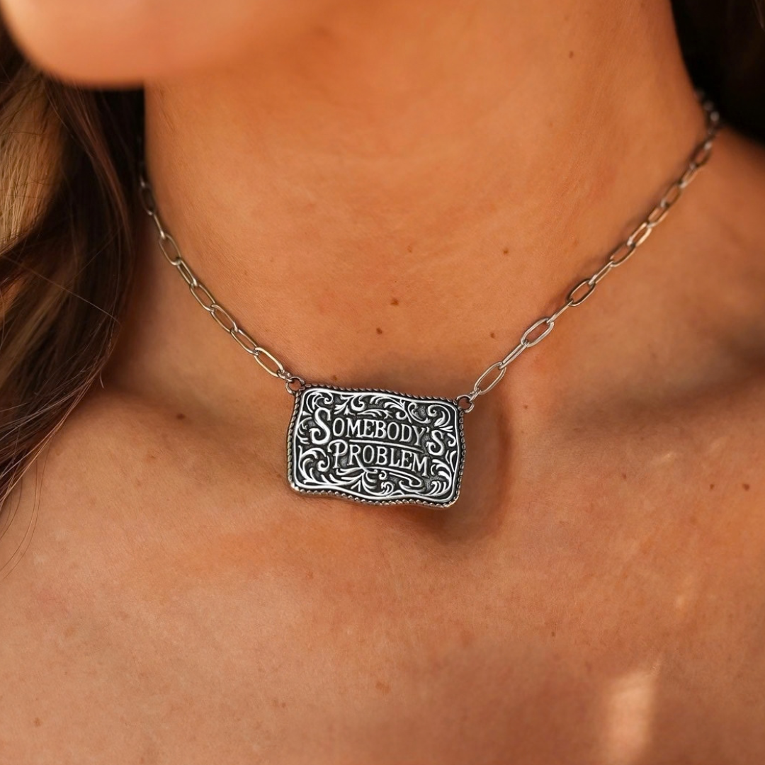 A silver-toned The Somebody’s Problem western statement necklace featuring a rectangular pendant with engraved scrollwork and the text SOMEBODYS PROBLEM.