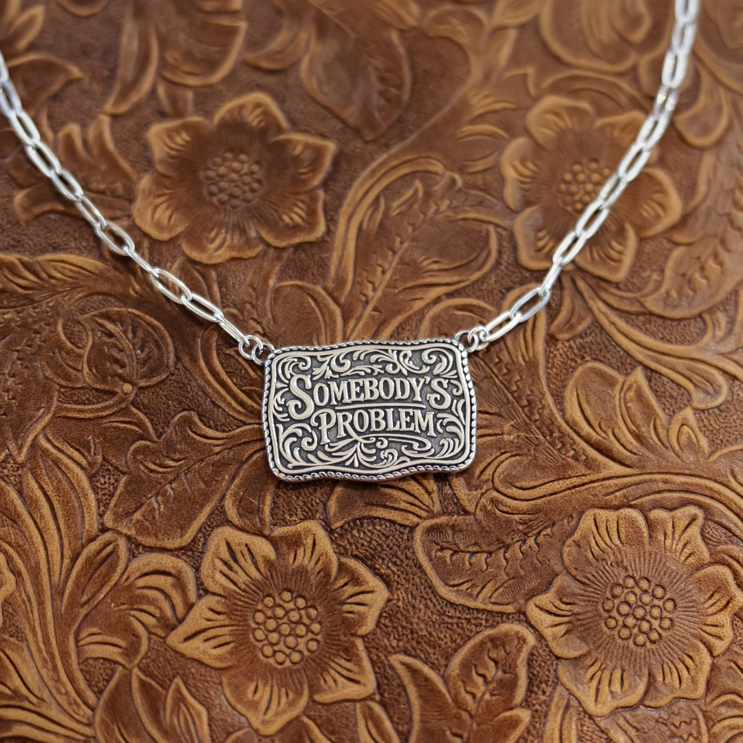 The Somebody’s Problem rectangular silver pendant on a chain, featuring Western-style engraving that reads Somebodys Problem surrounded by decorative scrollwork.