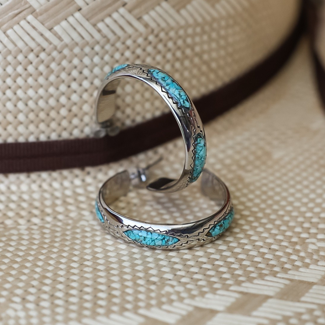 Two silver The Sunny Hoops earrings with turquoise inlay rest against a woven straw hat with a brown band. The hat’s textured pattern forms the background, highlighting these Western style earrings that capture the spirit of Southwestern jewelry.