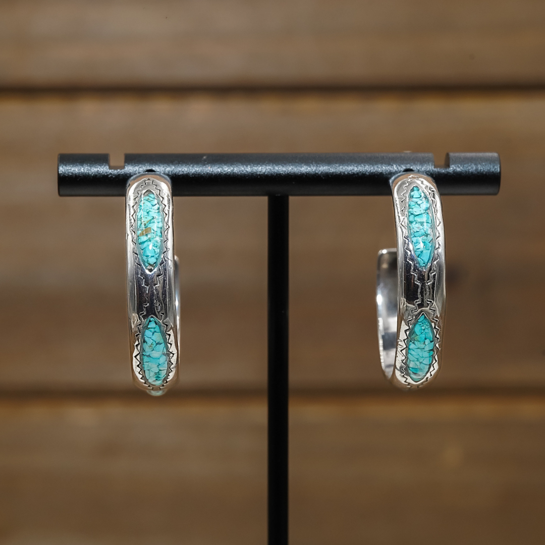 The Sunny Hoops capture the essence of Southwestern jewelry with their silver hoops featuring vibrant turquoise inlays and detailed engraving, beautifully displayed on a black stand against a blurred wooden background. These Western style earrings combine crushed turquoise accents with intricate designs for a truly striking look.