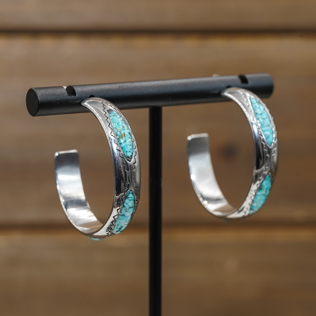 The Sunny Hoops are a pair of silver hoop earrings featuring crushed turquoise inlays and engraved patterns. Displayed on a black stand against a wooden background, these hoops embody the classic Southwestern jewelry style.