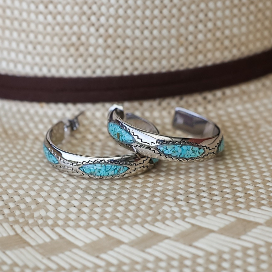 The Sunny Hoops, a pair of silver hoop earrings featuring turquoise inlay, rest atop a woven straw hat with a brown band, capturing the timeless allure of Southwestern jewelry.
