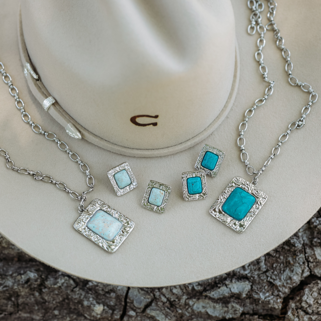 The Veryl—a striking statement silver necklace—along with three pairs of silver and turquoise or white stone earrings, are artfully arranged on a light beige cowboy hat atop a textured wooden surface, creating a classic display of Western jewelry.
