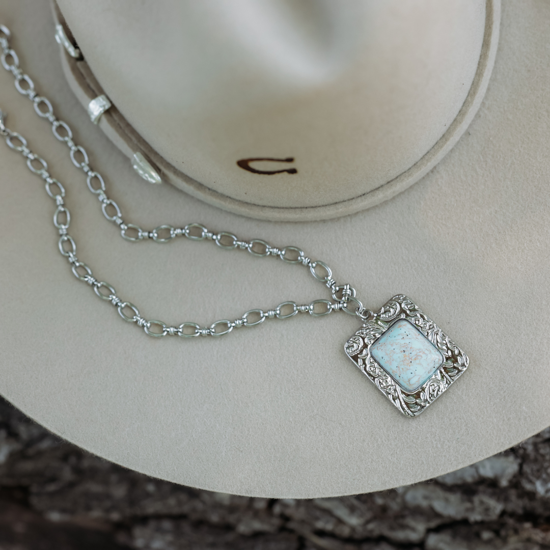 The Veryl, a silver statement necklace with a rectangular, ornate pendant featuring a light stone, rests atop a beige cowboy hat adorned with a horseshoe pin, capturing the timeless charm of Western jewelry.
