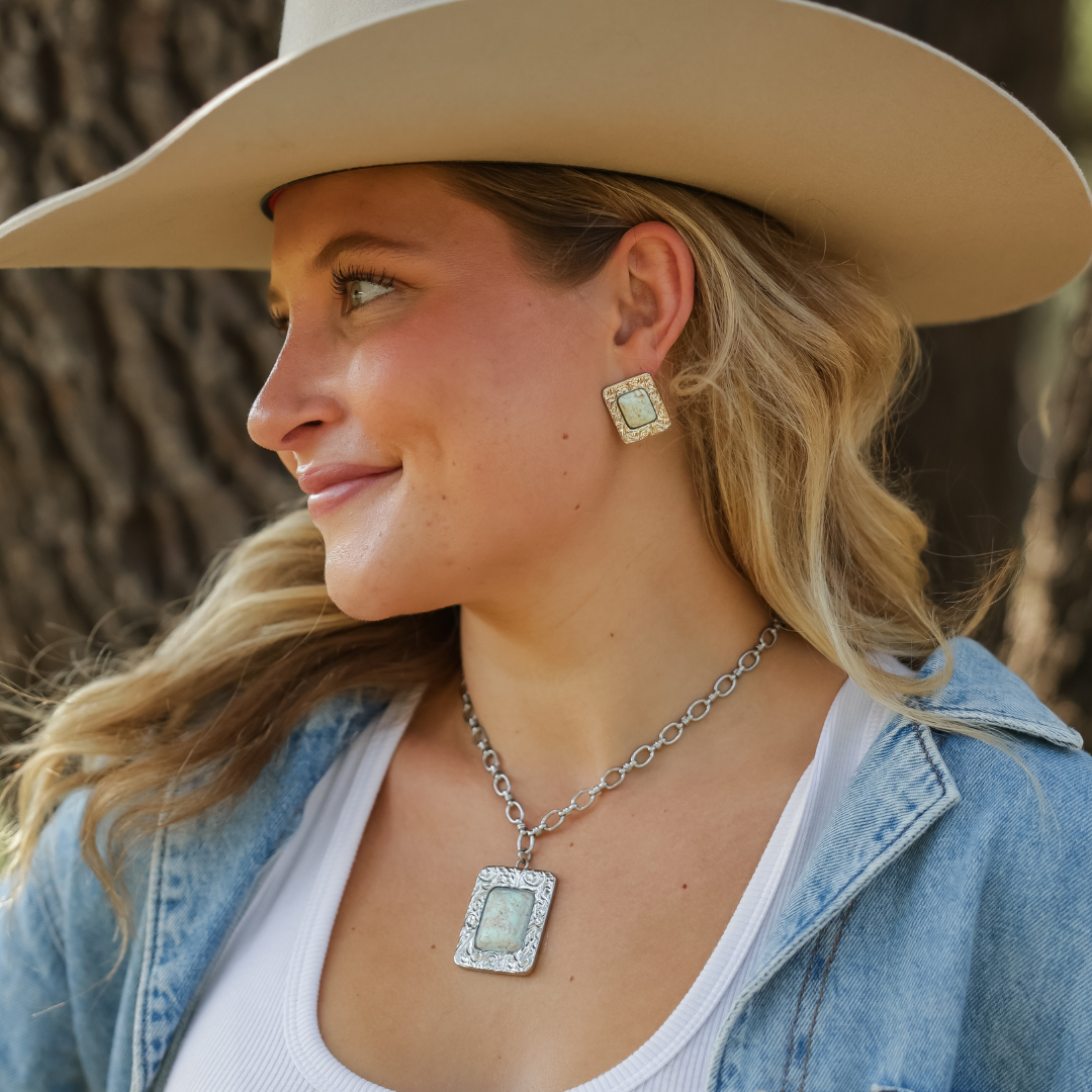 Smiling woman in a cowboy hat and denim jacket stands outdoors near a tree, wearing The Veryl—a matching silver statement necklace and earrings set with light green stones.