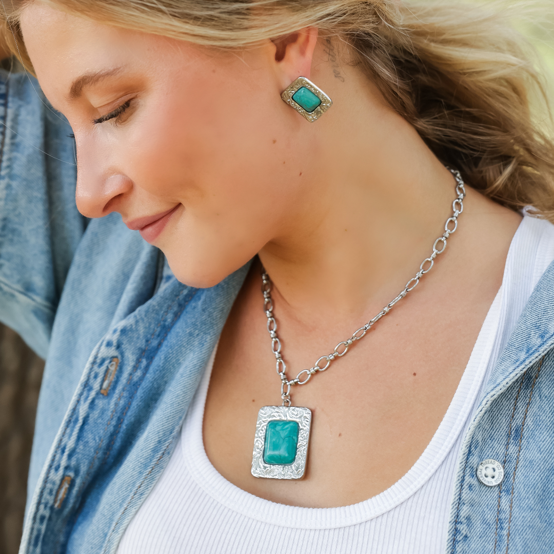 A woman in a denim jacket and white top wears The Veryl—a striking silver statement necklace and earrings set, both featuring large green stones—as she looks down and smiles outdoors.
