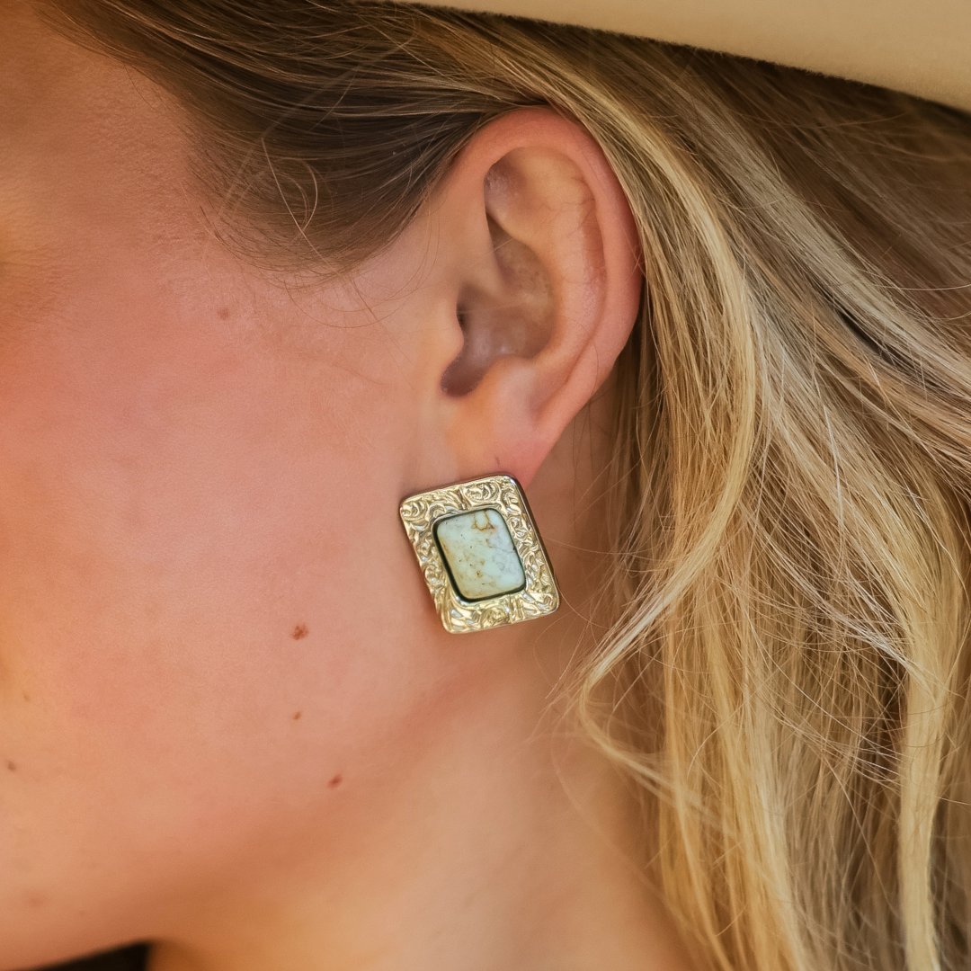 A close-up of a woman’s ear wearing The Veryl Stud Earrings—large, rectangular gold earrings featuring an ornate border and a light-colored center stone inspired by Western stud designs. She has blonde hair and is wearing a beige hat.