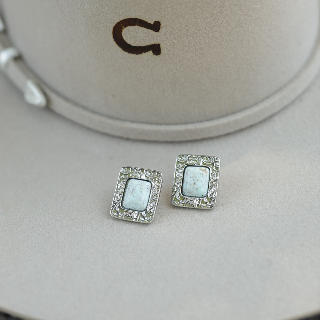 The Veryl Stud Earrings, featuring rectangular silver styling and white stone centers, rest on a light beige cowboy hat adorned with a dark horseshoe symbol.