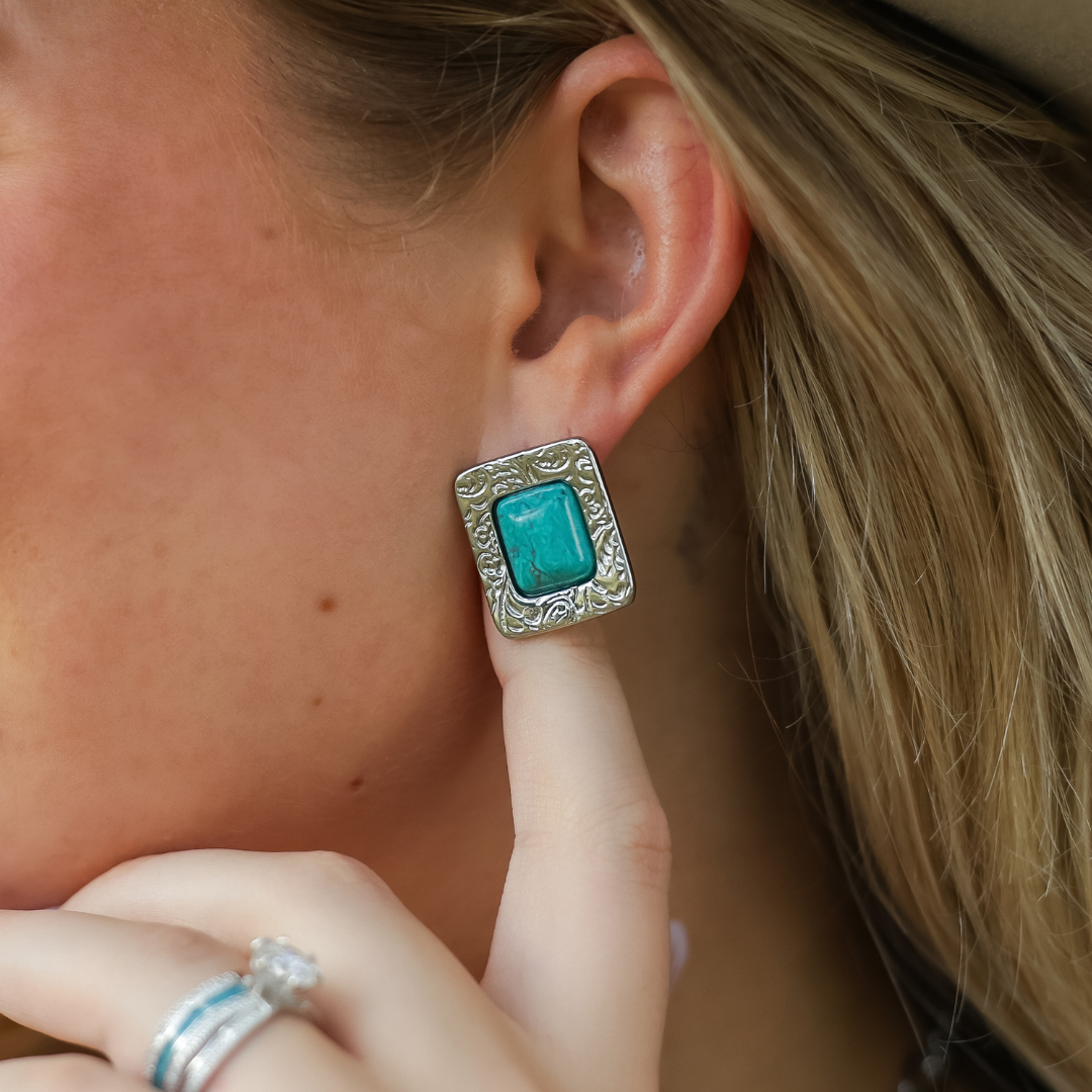 A close-up of a person wearing The Veryl Stud Earrings—a large, rectangular, silver turquoise design with engraved Western-inspired details. The person gently touches the earring while showcasing rings on their finger, embodying a bold style reminiscent of Veryl Goodnight.