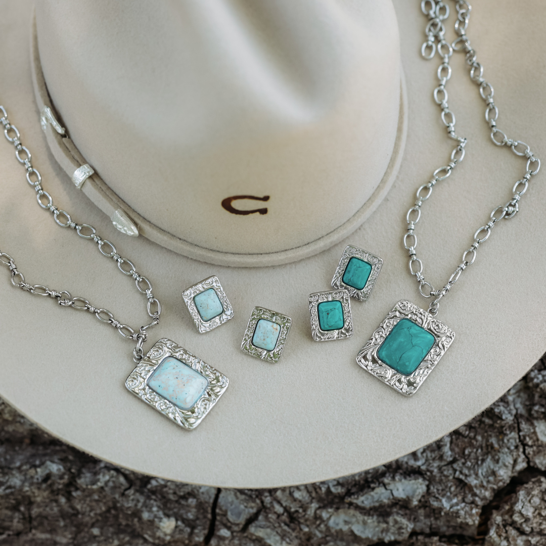 Two “The Veryl” silver necklaces and three pairs of matching earrings featuring turquoise and white stones are displayed on a beige cowboy hat and a textured surface, highlighting statement jewelry with bold Western necklace designs.
