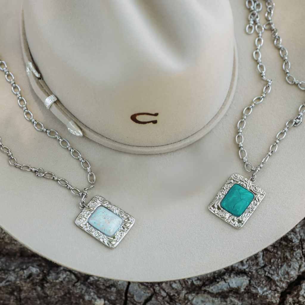Two silver chain necklaces from The Veryl collection, each featuring a square gemstone pendant—one in white and the other in turquoise—are displayed atop a beige cowboy hat with a silver band, set on a rough textured surface. Perfect for bringing Western jewelry charm to your look, these necklaces make an effortless statement duo for any occasion.