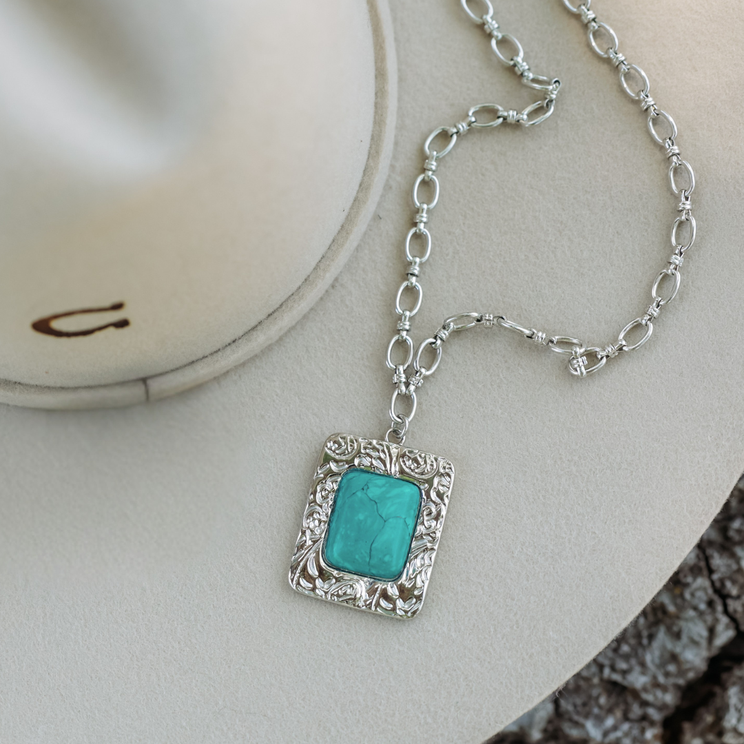 The Veryl, a silver turquoise necklace with a rectangular pendant, rests on a light beige cowboy hat partially visible beside rough tree bark, embodying the bold spirit of statement jewelry and Western charm.
