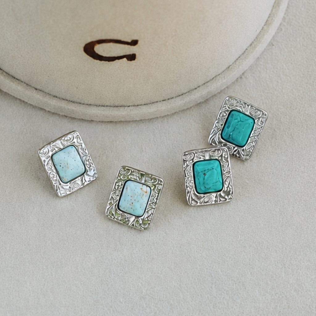 Four silver stud earrings with ornate edges, each featuring a square turquoise or light blue stone reminiscent of classic Western style, are displayed on a beige surface near a suede jewelry box with a brown C embroidered on it. Product Name: The Veryl Stud Earrings.