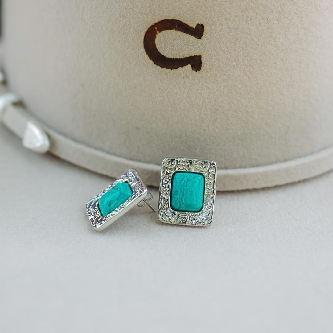 The Veryl Stud Earrings—two silver Western studs featuring intricate designs and rectangular turquoise stones—are displayed on a light beige surface in front of a cream-colored hat adorned with a brown C logo, evoking the timeless elegance characteristic of Veryl Goodnight’s artwork.