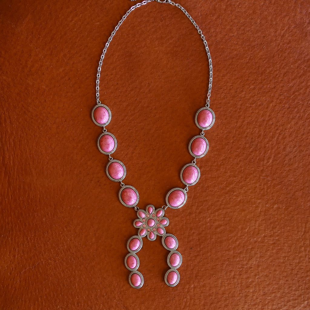 The Charlie is a silver squash blossom necklace featuring oval pink rhodonite stones arranged in a symmetrical pattern, with a central flower-shaped cluster and two curved dangles, shown against a brown textured background.