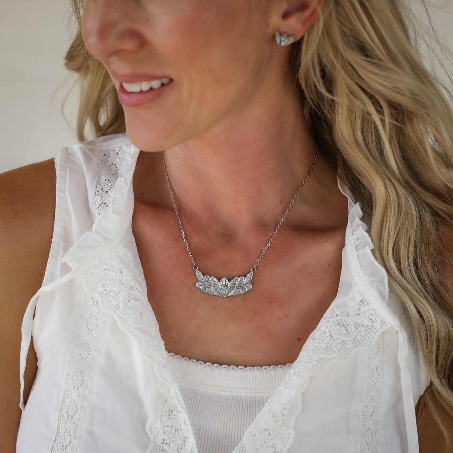 Woman with long blonde hair wearing a white sleeveless blouse, silver earrings, and The Dessie—a detailed Western necklace featuring a leaf design—smiling and gazing to the side against a light background.
